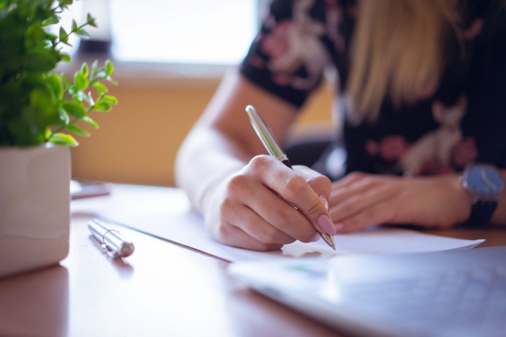 Close-up of woman's hand writing with pen at office desk on a sunny day, plant decor nearby.