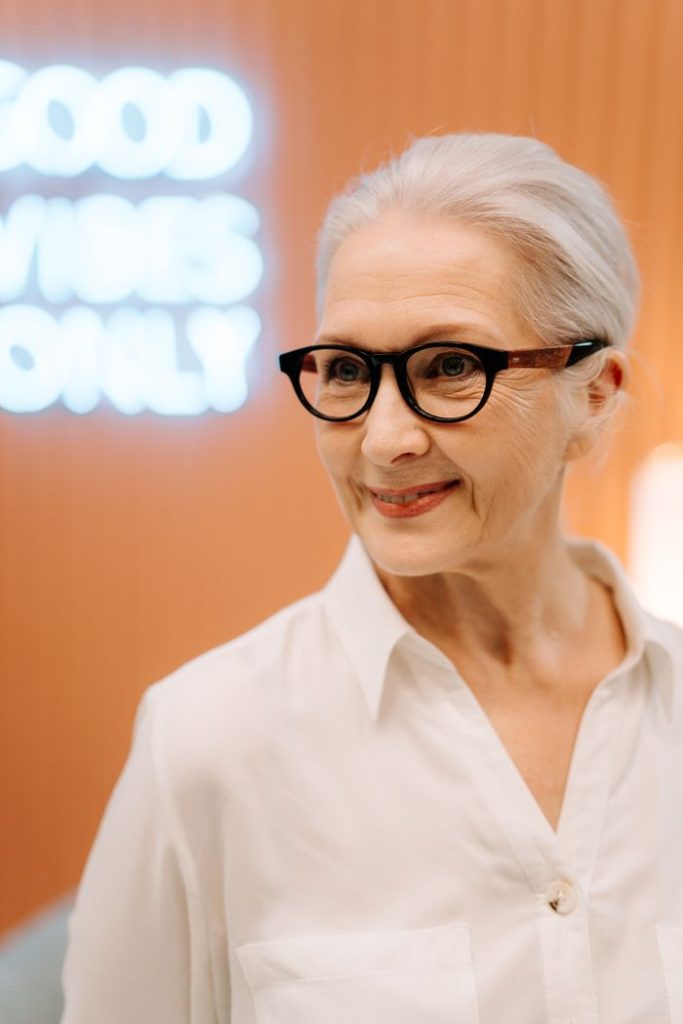 La paradoja de la posmenopausia: por qué la etapa más larga de tu vida sigue siendo la más olvidada Elderly woman smiling brightly with eyeglasses against a warm indoor backdrop.