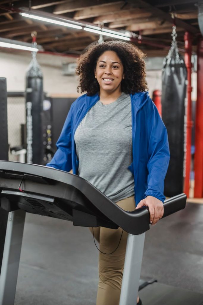Smiling plus size black woman walking on treadmill while cardio exercising in modern gym