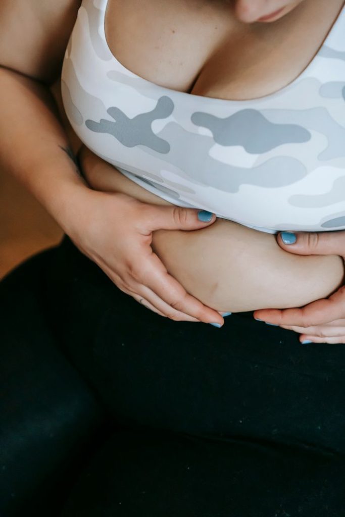 From above of crop anonymous obese female in bra and leggings sitting and touching stomach fat after weight lose workout in gym