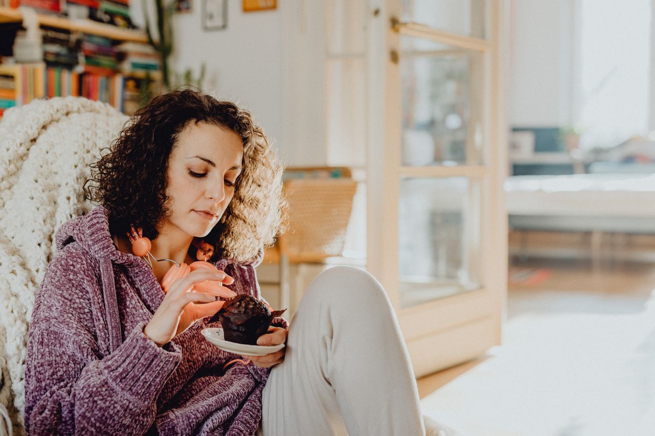 Woman with curly hair enjoying a cupcake in a cozy living room setting.