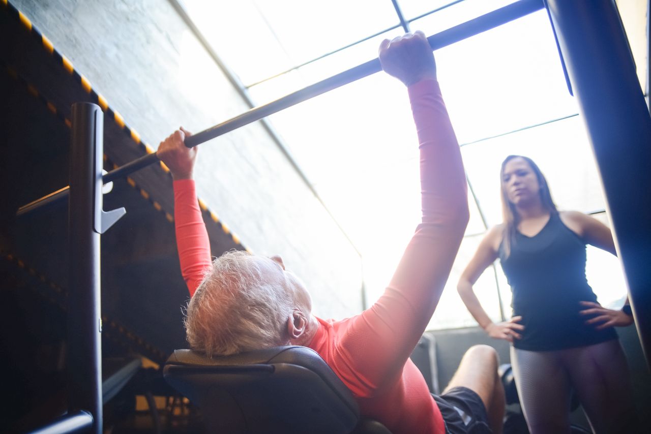 Senior man bench pressing with a female trainer overseeing his workout in an indoor gym.
