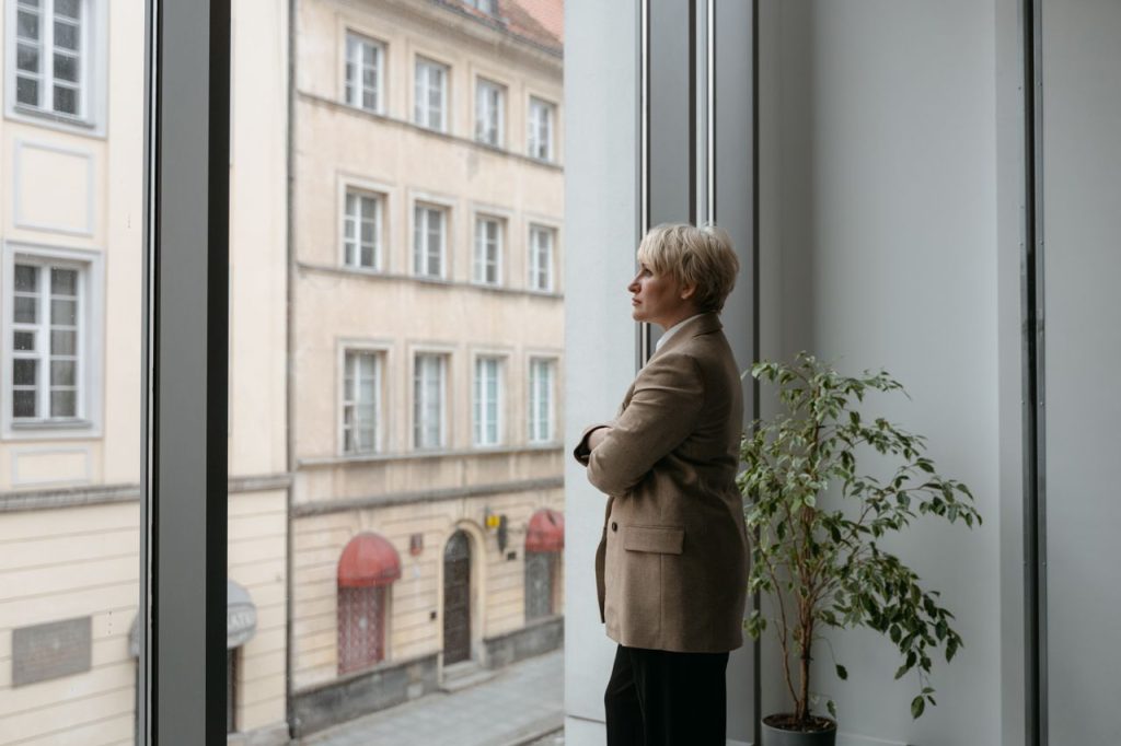 A thoughtful woman stands by a large window watching the cityscape outside, arms crossed