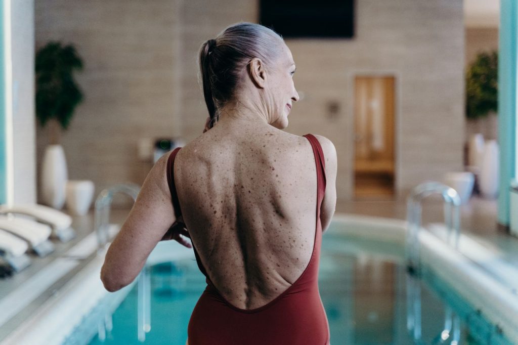 La paradoja de la posmenopausia: por qué la etapa más larga de tu vida sigue siendo la más olvidada Senior woman in red swimsuit standing at indoor pool side, exuding confidence.