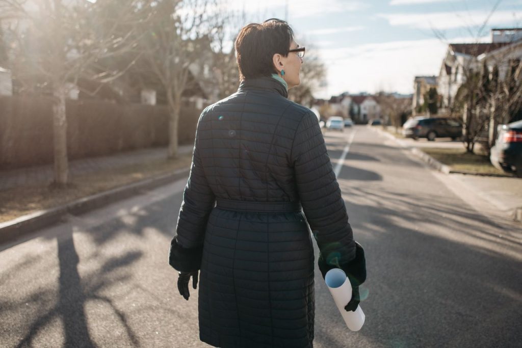 Woman in blue coat holding paper roll on sunny residential street.