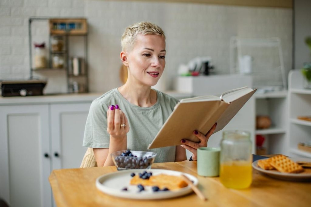 Mujer en cocina con libro Mujer rubia de pelo corto, de 40 años, atractiva, en la cocina, leyendo mientras come unos arándanos